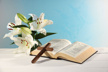 Wooden cross, Bible and beautiful lily flowers on white table against light blue background....