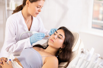 Young brunette undergoing device-driven facial treatment using ion bubble pen attachment for gentle exfoliation, deep hydration and skin revitalization at cosmetic medicine clinic