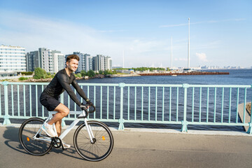 A man cycles along a waterfront path on a bright day, with a backdrop of modern buildings and a...