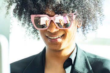 Young non-binary mixed-race person with an afro and sunglasses, wearing a black business suit. Smiling while a reflection is seen in the glasses.