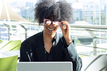 Non-binary young person with afro hair and sunglasses, in a black business suit, holding the sunglasses while smiling and looking at the laptop.