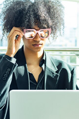 Young non-binary person with an afro and sunglasses, wearing a black business suit. Working with their white laptop.