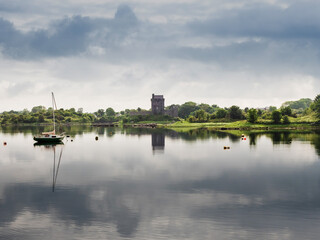 Obraz premium Small sailing boat and Dungauaire castle in the background with sky reflection in calm water surface. Relaxing mood. Popular landmark in Kinvarra town, Ireland. Irish nature scene.