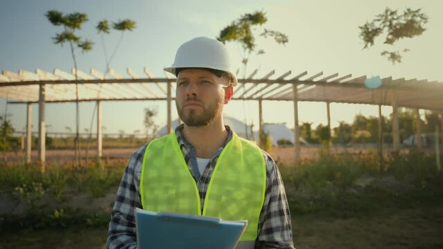 Caucasian millennial 30s successful male engineer man guy hardhat specialist technician worker builder inspector holding tablet pen writing quality report standing outside farmland field construction