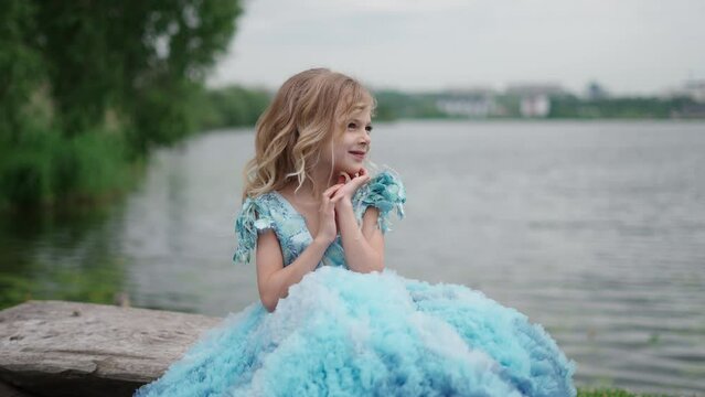 Little girl in blue dress is sitting on shore of lake. Little princess concept.
