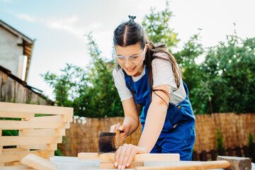 One young caucasian carpenter woman is using sawdust and hammer making things from wood and planks in her backyard during the day	