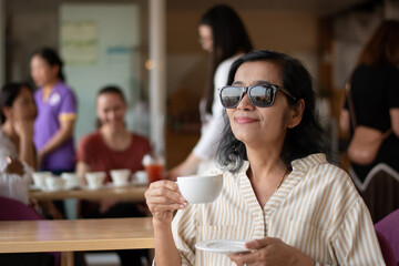 Asian mid adult woman sits in the middle of the cafe and drinking coffee, soft focus, blurred people in cafe background.