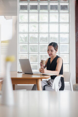 A young professional woman working on her laptop at a modern cafe, holding a coffee cup and smiling, with a glass block wall in the background.