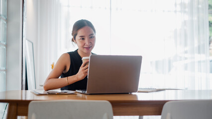 Fototapeta premium A young woman working on her laptop in a bright home office, holding a coffee cup. Modern workspace with natural light.