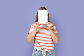 Portrait of shy anonymous blond woman wearing striped T-shirt holding organizer with empty paper in front her face, copy space for advertisement. Indoor studio shot isolated on purple background.