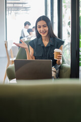 A young woman holding a coffee cup, smiling and engaged in a video call while working on her laptop in a modern cafe with natural light.