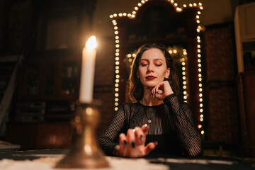 Naklejka premium Portrait of enigmatic fortuneteller dealing Tarot cards forecasting future for client by light of burning candles. Fortune-telling having divination session with tarot cards at dark esoteric room.