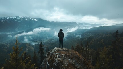 A figure stands atop a windy mountaintop facing away from the camera but still taking in the stunning view of sprawling forests and . .