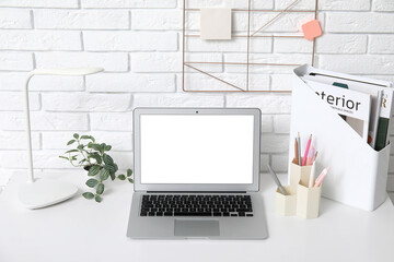 Modern workplace with laptop, lamp, magazines and stationery near white brick wall