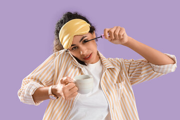 Hurrying young African-American woman in pajamas with coffee cup and mobile phone applying mascara...