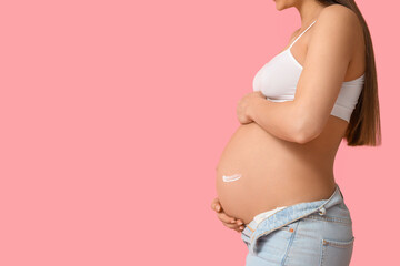 Young pregnant woman applying cream against stretch marks on pink background