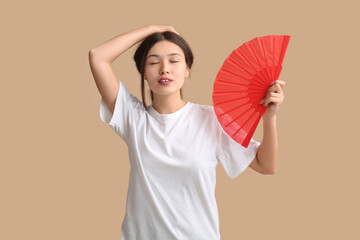 Young woman with hand fan suffering from heat on beige background