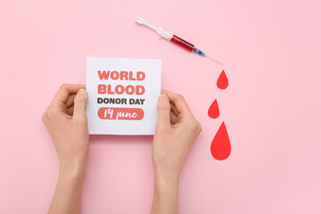 Female hands holding paper sheet with sign WORLD BLOOD DONOR DAY 14 JUNE, paper blood drops and syringe on pink background