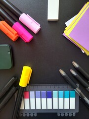 Black desk with various colorful writing utensils. There are orange, pink, and light pink markers, a yellow highlighter, a black pen, and a white eraser, colorful sticky notes.