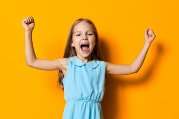 Joyful little girl in a blue dress raising her arms in excitement on a vibrant orange background