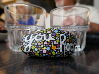 You rock kindness rock with drinking glasses and people out of focus in background