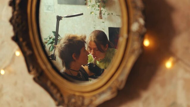 Mirror reflection of female tattoo artist in gloves using needle as doing upper arm tattoo for young woman in home studio