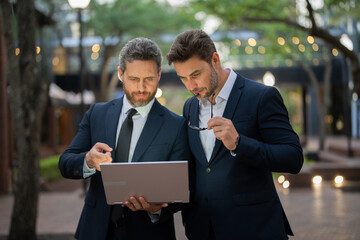 Two businessmen on the street, deeply engrossed, eyes fixed on their laptop screen, perhaps...
