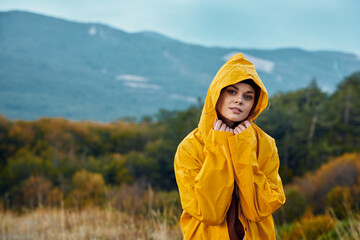 Woman in yellow raincoat standing in picturesque field with majestic mountains in background, outdoor travel adventure beauty scene