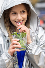 A young woman in a silver hooded jacket happily drinking a mint beverage outdoors, embodying a...