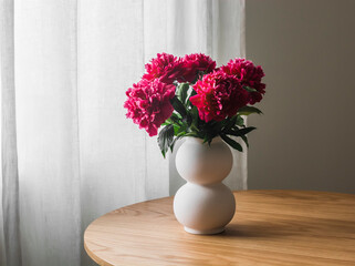 A bouquet of red peonies in a ceramic vase on a round wooden table in the living room