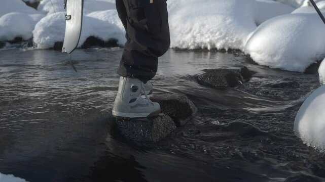 A person steps on rocks crossing a snowy stream in Hokkaido, Japan, carrying a splitboard