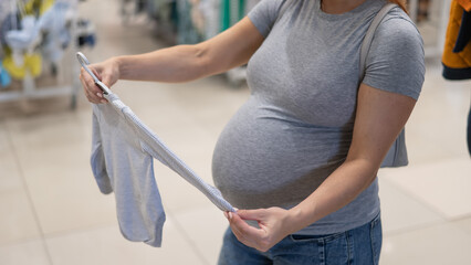 Caucasian pregnant woman chooses baby clothes in a store. Faceless expectant mother in the 3rd trimester. 