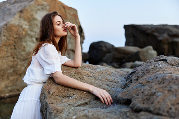 Serene woman in white dress sitting on ocean rock with hand on face gazing into distance