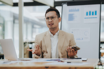 Cheerful young Asian businessman smiling in bright modern office working on laptop computer checking charts, graphs and paper.