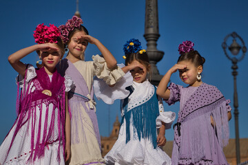 Naklejka premium Four girls dancing flamenco, covering their eyes from the sun with their hands, in typical flamenco costumes on a bridge in a beautiful square in Seville. Dance concept, flamenco, typical Spanish.