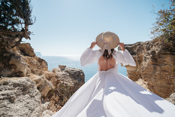 A woman in a white dress is standing on a rocky cliff overlooking the ocean. She is wearing a straw...