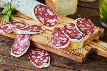 Sliced dried or smoked sausage with white mold on a wooden table. There is a loaf of bread in the background. Meat production.