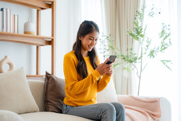 Obraz premium Happy young asian woman relax on comfortable couch at home texting messaging on smartphone, smiling girl use cellphone chatting, browse wireless internet on gadget, shopping online from home