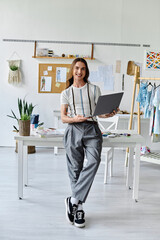 A young man stands in his clothing restoration atelier, laptop in hand, showcasing his passion for sustainable fashion and giving new life to discarded clothing.