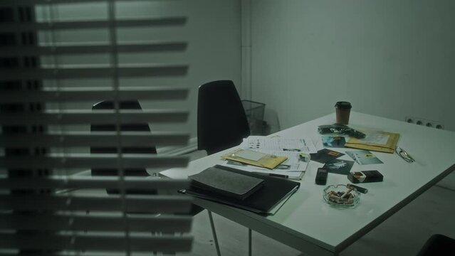 Full no people shot from window blinds of empty interrogation room at police department, table with evidence envelopes, crime scene photos, gun in plastic bag, coffee cup, full ashtray with cigarette