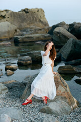 Serene woman in white dress sitting on seaside rock with vibrant red shoes by ocean view