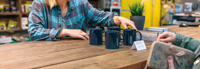 Banner of unrecognizable woman employee showing blue mugs to customer in vintage shop. Female client holding her wallet to pay purchase while happy cashier offering discount cups on local store.