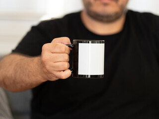 A man in a black t-shirt holding a black mug with a white label. Mug to showcase your design. Cropped close-up image