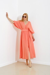A stylish woman in a coral maxi dress with a v-neckline leaning against white wall