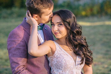 Stylish groom hugs beautiful bride from behind in nature, standing on green grass in park, garden.
