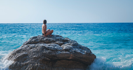 A young woman in a bikini sits on a large stone on the beach and looks at the sea, relaxing and...