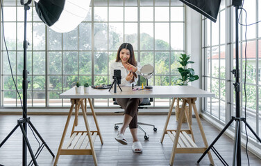 A woman filming her makeup tutorial in a bright room with large window, using a studio setup that include a smartphone mounted on a tripod, lighting equipment and various makeup product on the table.