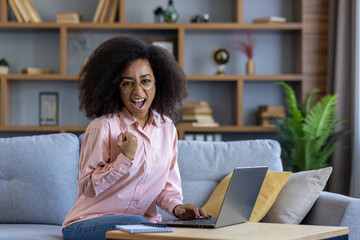Happy woman celebrates success while working on a laptop at home. Experience joy and accomplishment in a cozy living space.