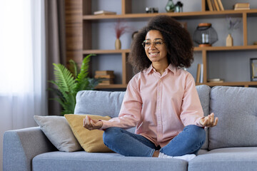 Smiling woman practicing yoga and meditation on a cozy sofa at home. Relaxed, comfortable, and content in a peaceful indoor setting.