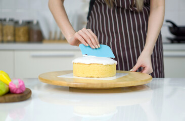 Closeup hand holding plastic cake cream spatula, spread the top of the cake with white cream,...